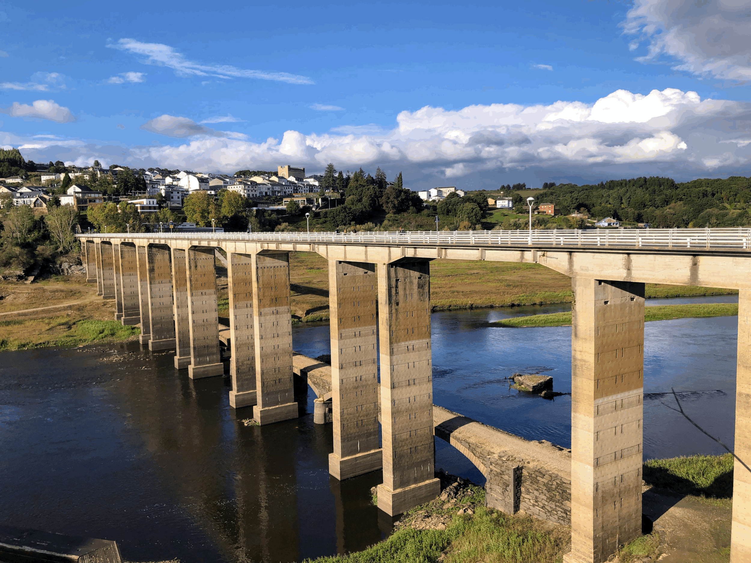 A concrete bridge spanning over a river with a hillside neighborhood in the background under a partly cloudy sky.