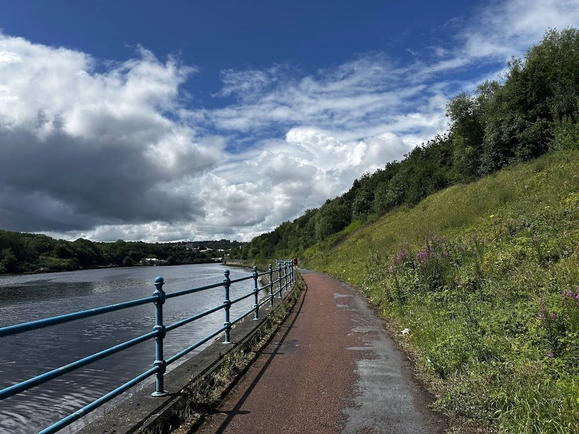 riverside asphalt trail with railing and green countryside and cloudy but blue skies