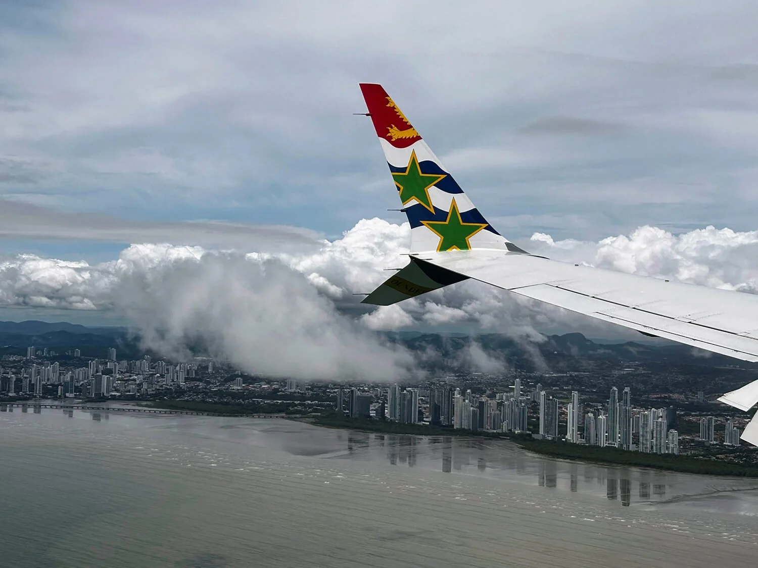 View from an airplane window showing the airplane's wing with colorful design and a city below, with cloudy sky and mountains in the distance.