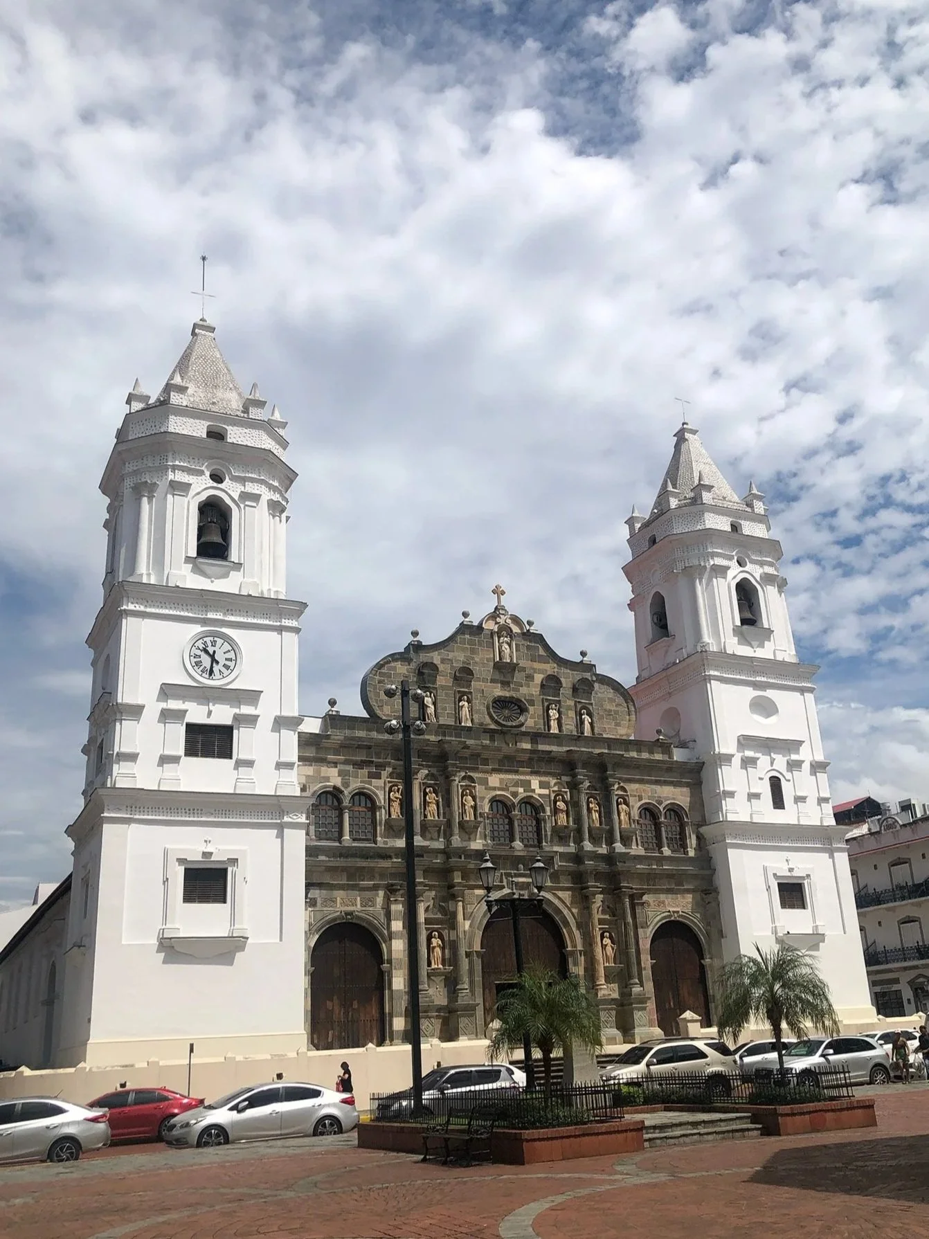 Front view of a historic church with two white bell towers, a clock on the left tower, and a decorative facade with statues and intricate details, under a partly cloudy sky.