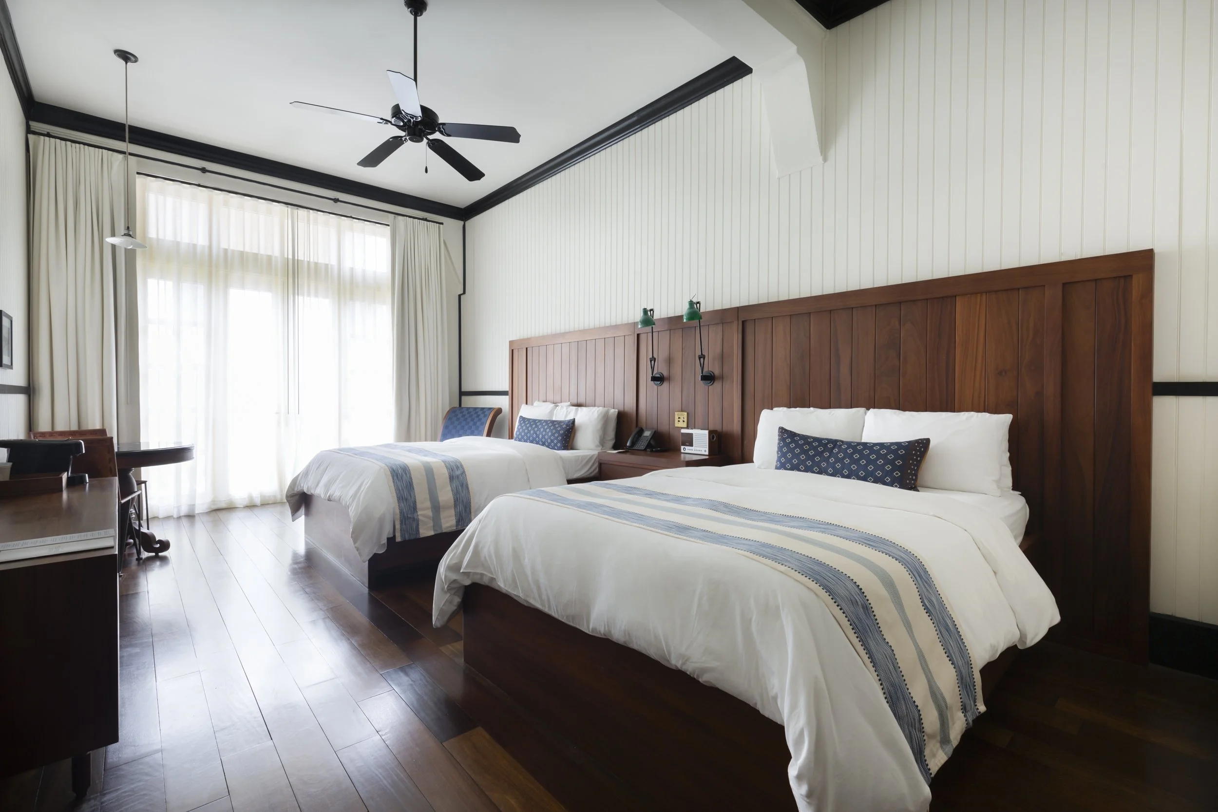 Two double beds with white linens and blue striped runners, wooden headboards, bedside lamps, and a nightstand in a hotel room with large windows and white curtains, ceiling fan, and wood flooring.