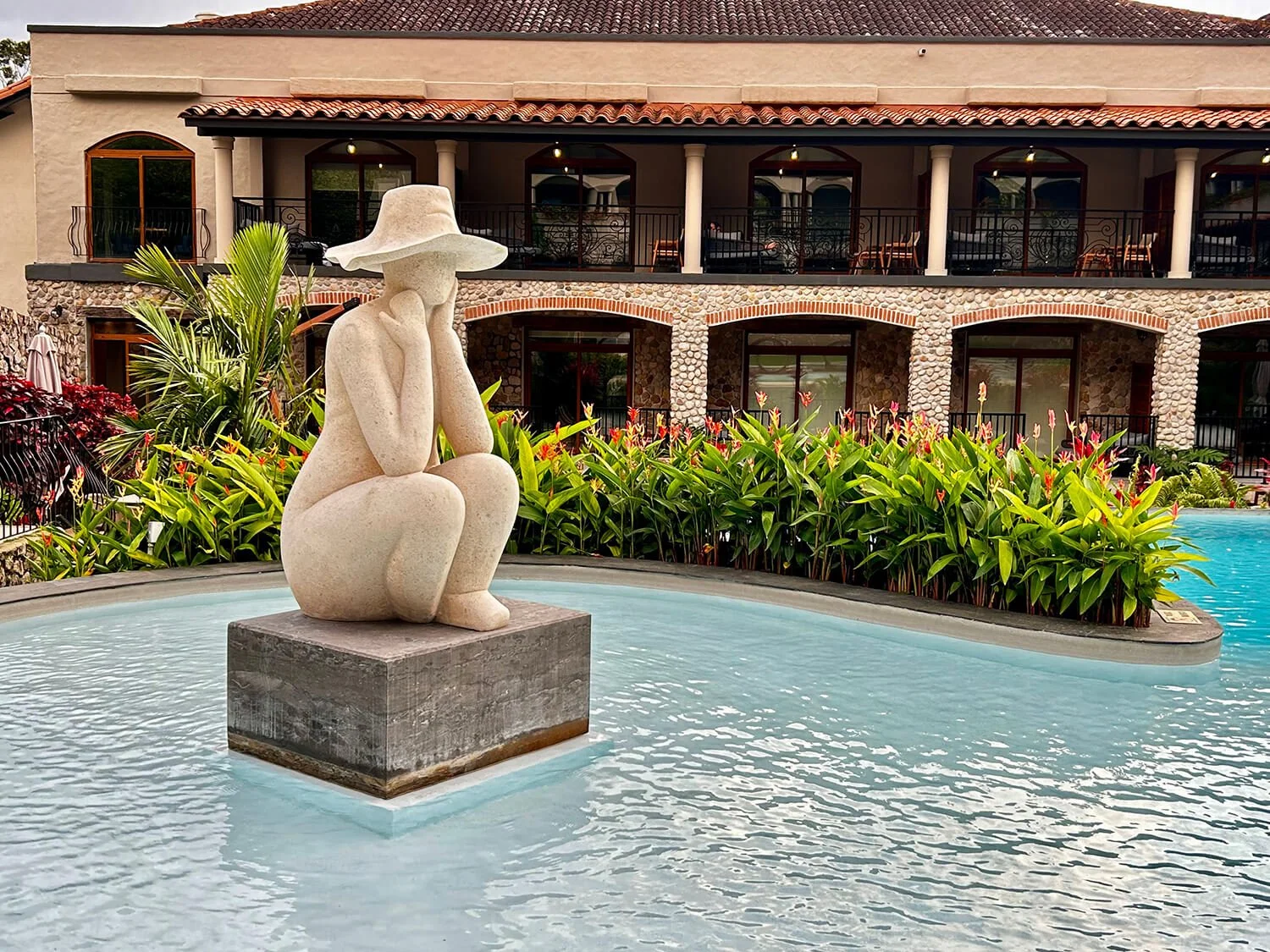 Statue of a woman sitting with her chin resting on her hand in a pool, surrounded by tropical plants with a building in the background.