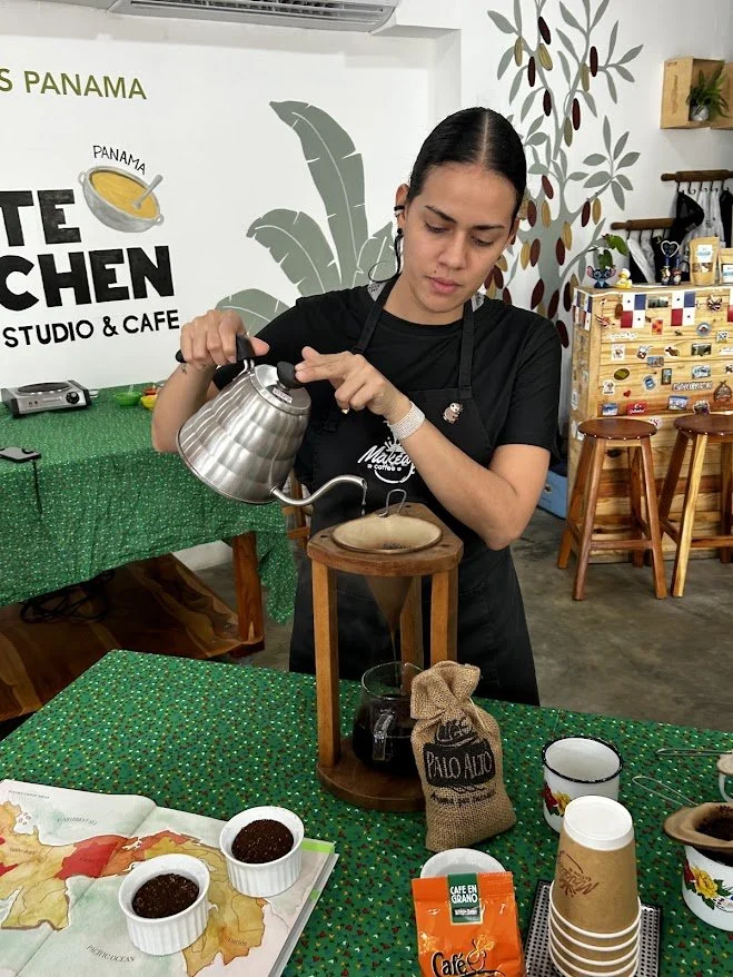 Woman in black shirt pouring hot water from a metal kettle into a coffee dripper on a wooden stand, surrounded by cups, coffee packets, and a map, in a cafe decorated with plants and a mural.