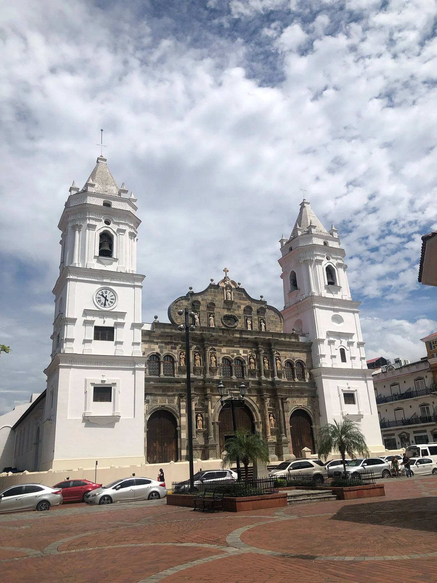 A historic church with two white bell towers, a detailed facade with statues, and a clock on one tower, set in an urban square with parked cars and a few pedestrians, under a partly cloudy sky.
