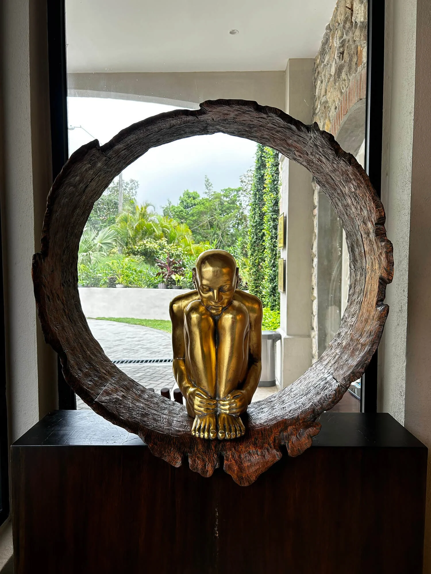 A gold sculpture of a seated person with knees drawn up and head bowed, placed inside a circular, wooden frame with a natural, textured finish, looking out to a lush outdoor scene.
