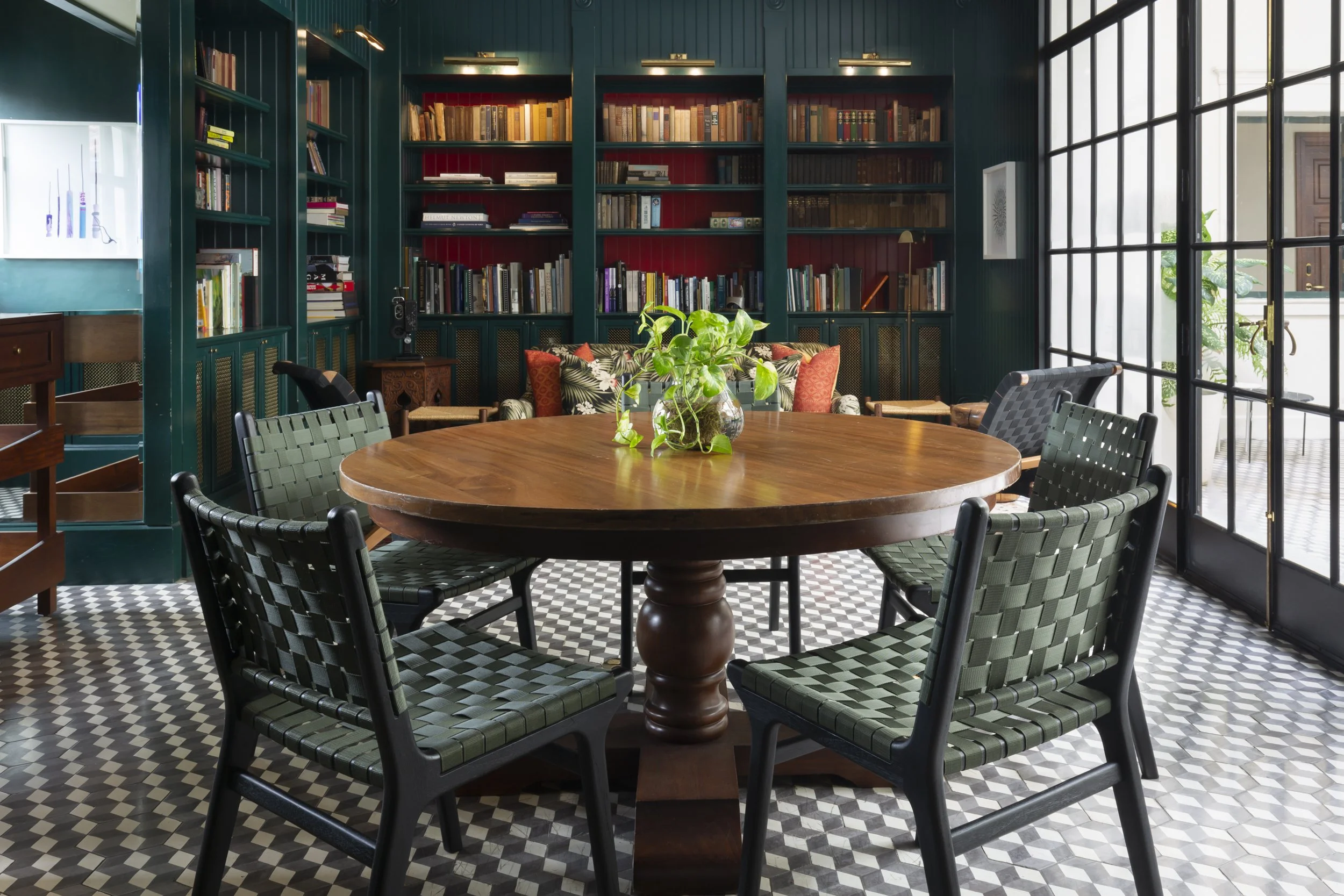 A cozy reading room with a wooden round table surrounded by six green patterned chairs, a large built-in bookshelf filled with books on the wall behind, and large glass doors letting in natural light, with a potted plant in the center of the table.