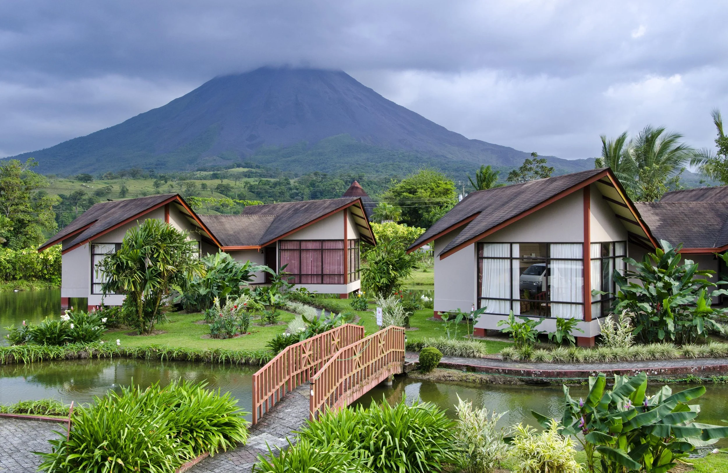 A scenic view of a volcano in the background, with lush greenery and a few buildings with traditional roofs and large windows in the foreground, along with a small pond and a decorative bridge.
