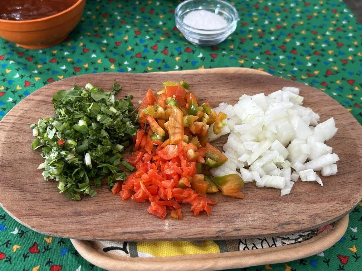 Chopped green onions, diced tomatoes, sliced jalapenos, and chopped onions on a wooden serving platter, with bowls of salt and a sauce in the background on a colorful, patterned tablecloth.