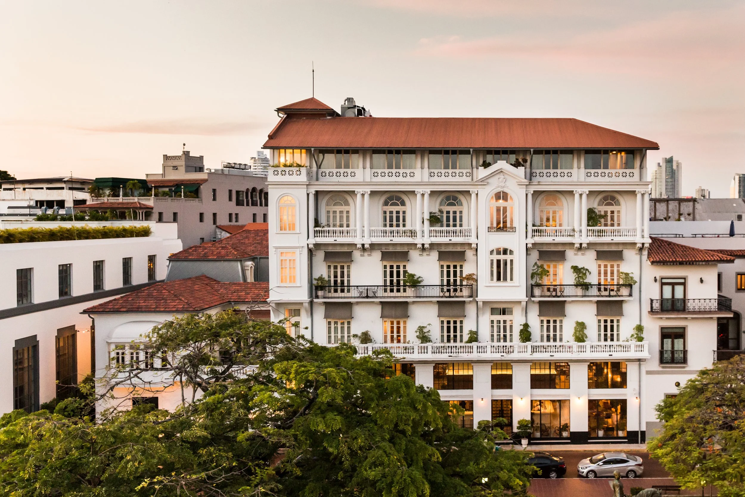 A multi-story white building with classical architectural details, including columns and balconies, is surrounded by trees and parked cars during dusk.