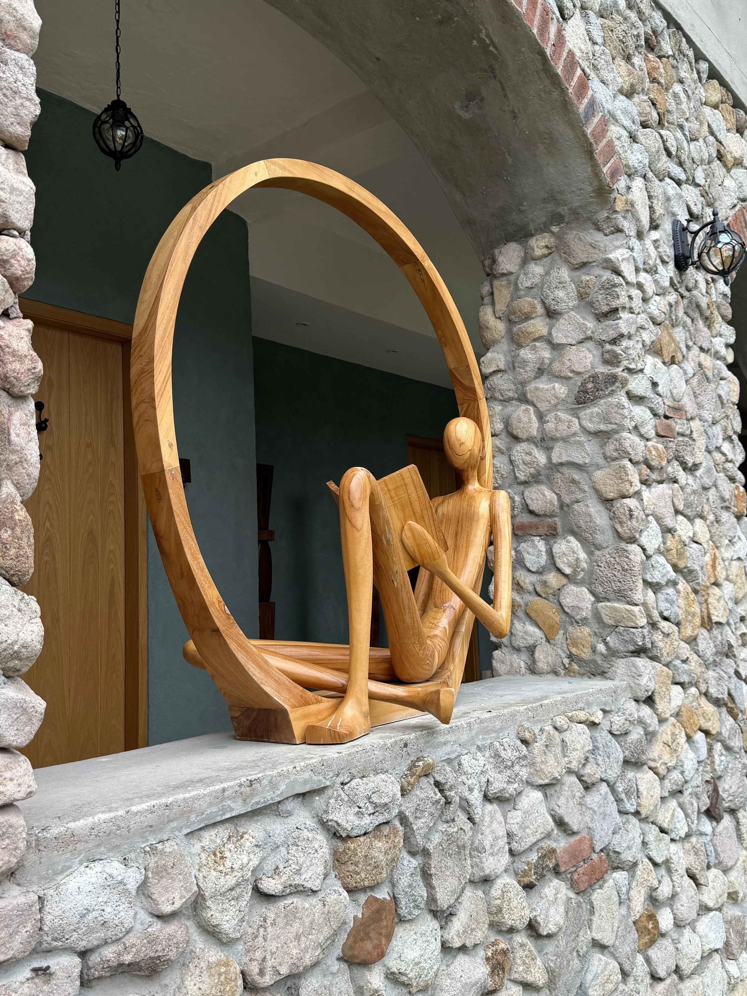 A wooden chair with a backrest that resembles a human figure is placed in a niche with a stone and brick wall. There are two black outdoor wall lanterns and a hanging lantern visible in the space.