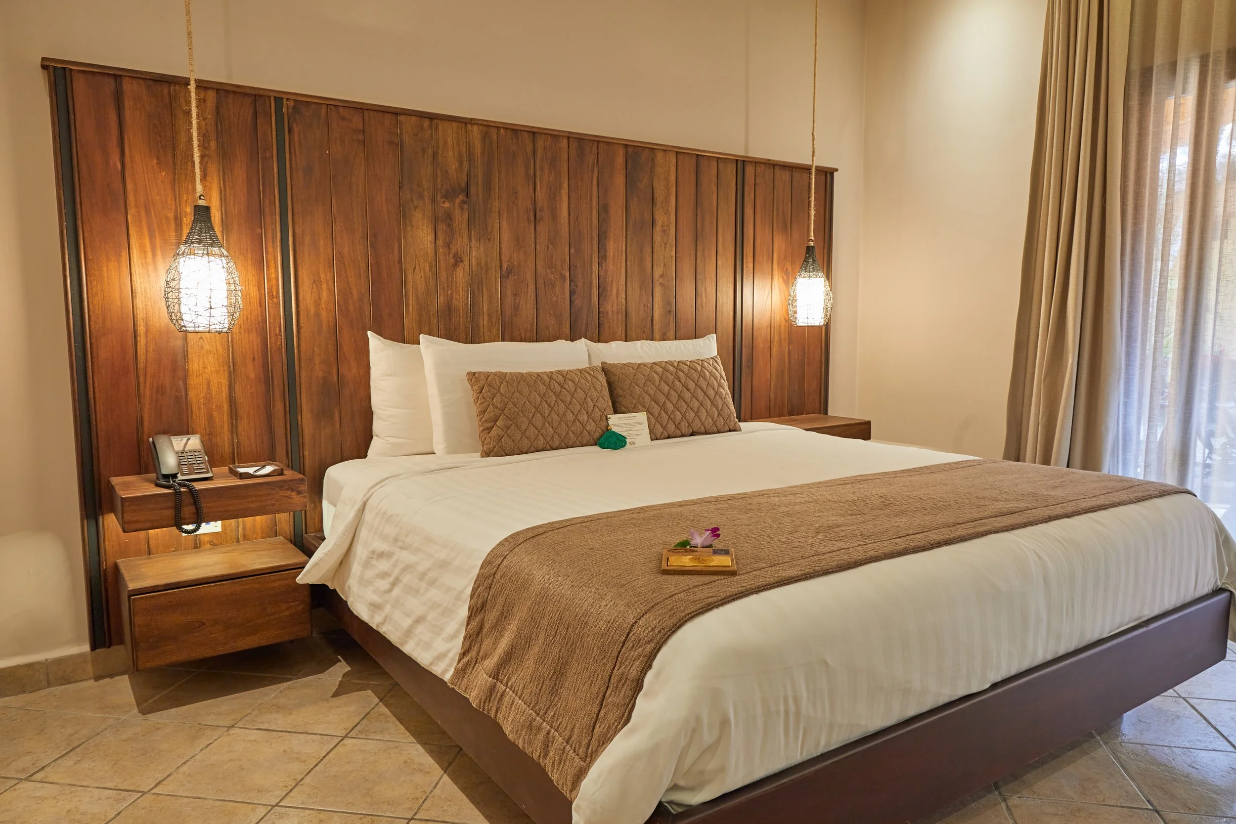 A hotel room with a large bed featuring white linens and brown accent pillows. There is a wooden headboard and matching bedside table with a telephone and a notepad. Two pendant lights hang on either side of the bed, and a window with curtains is on the right side, allowing natural light into the room.