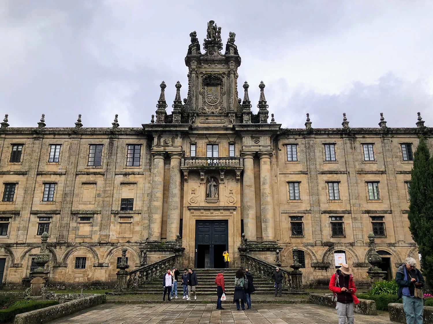 An old historic stone building with steps leading up to the entrance, people gathered in front, cloudy sky above, trees on sides.