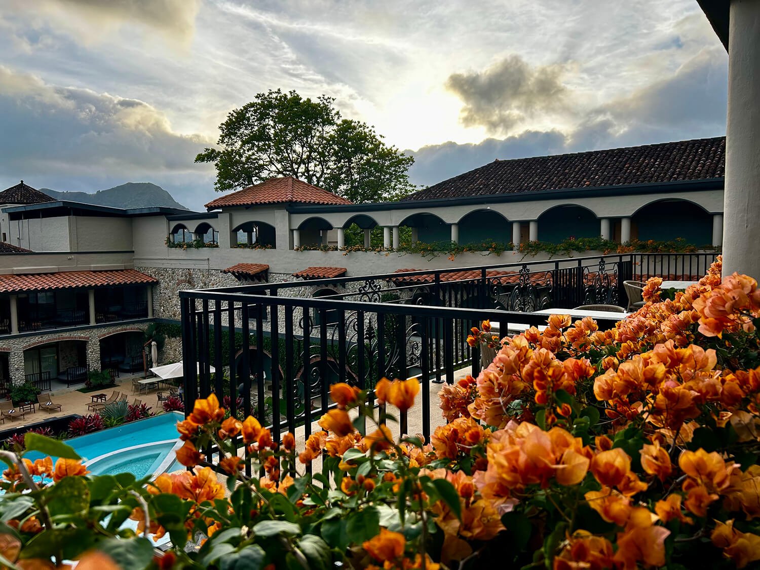 View of a residential or hotel courtyard with a swimming pool, outdoor chairs, and colorful flowers, overlooking a multi-story building with arched balconies and a scenic mountain background at sunset.