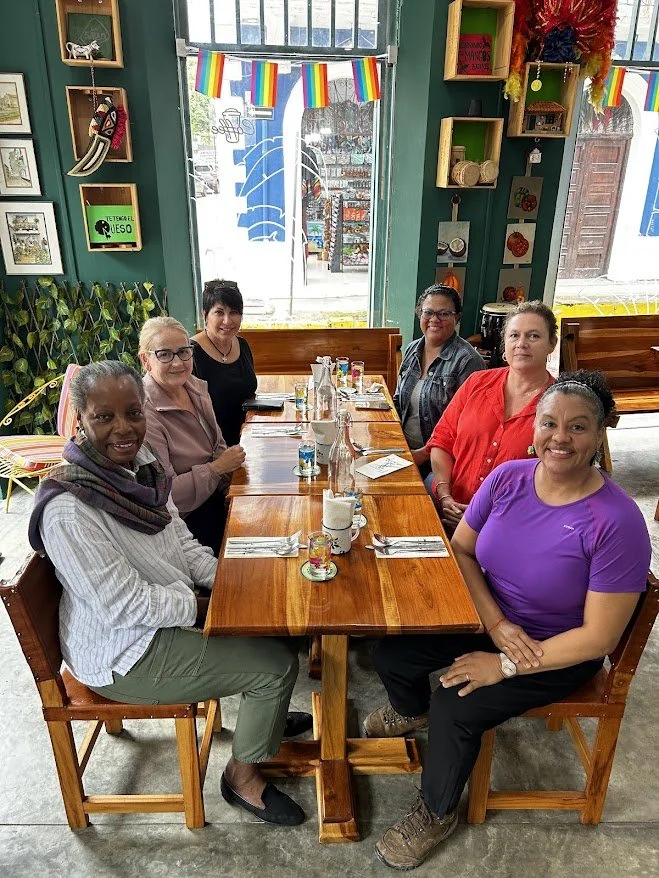 Six women sitting around a wooden table in a brightly decorated restaurant, with a window and colorful decorations in the background.