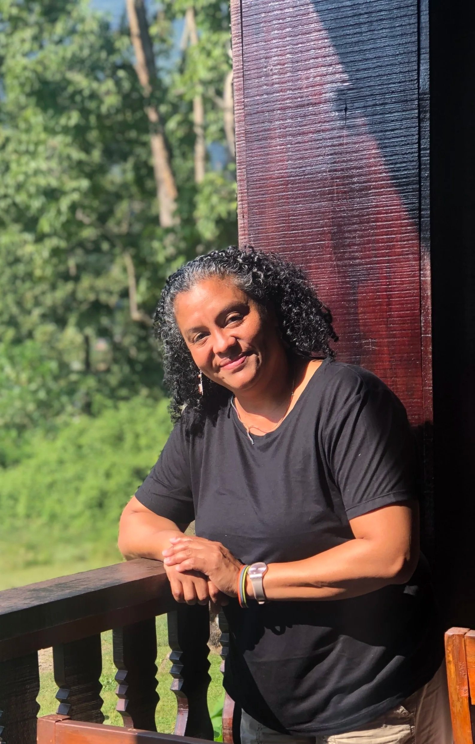 A woman with curly dark hair, wearing a black shirt and a rainbow bracelet, standing outdoors on a wooden balcony, smiling at the camera with a green, wooded background.