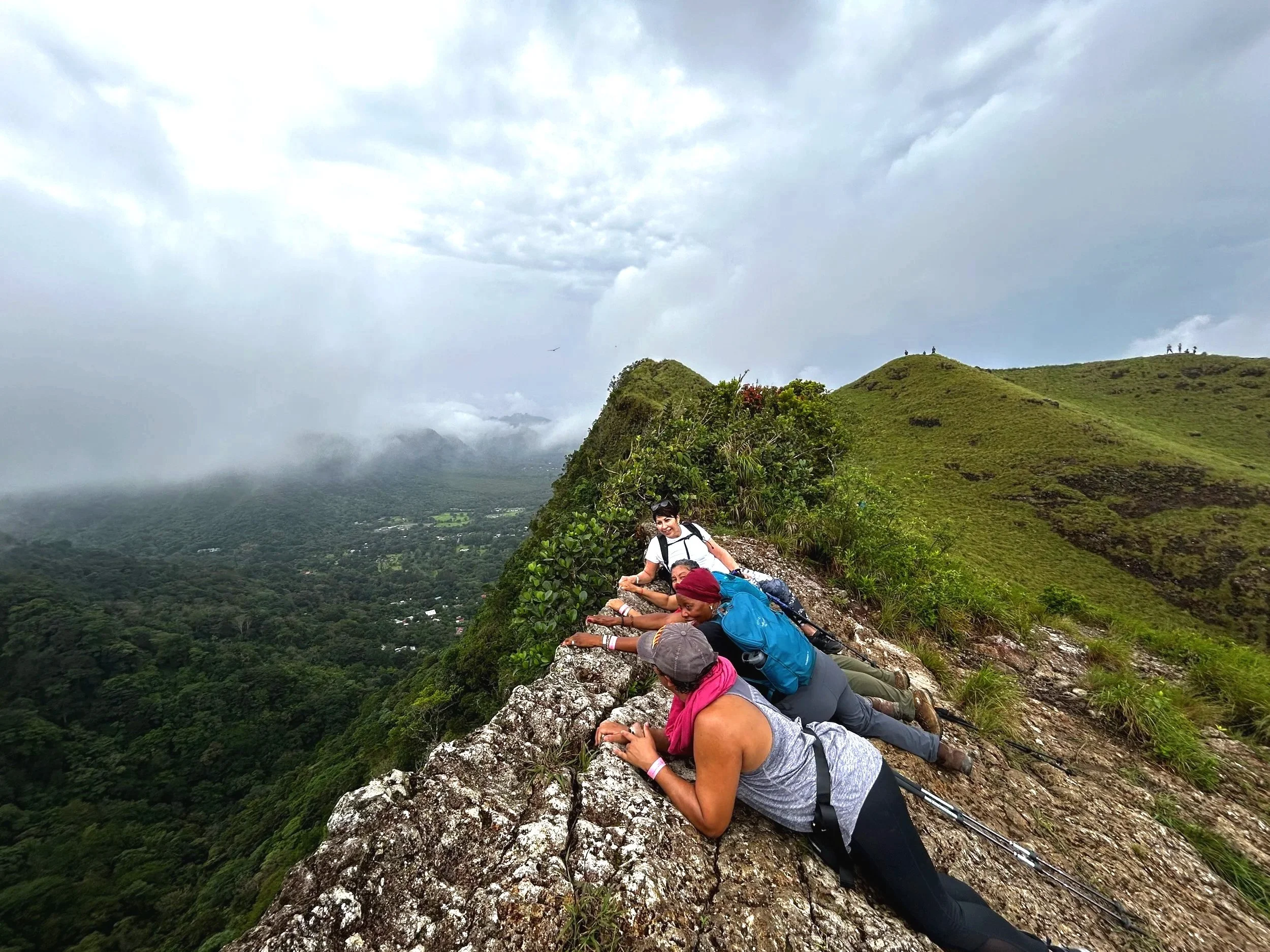A group of people lying and leaning on a rocky ledge along a mountain trail with a lush green valley below and rolling hills in the distance under a cloudy sky.