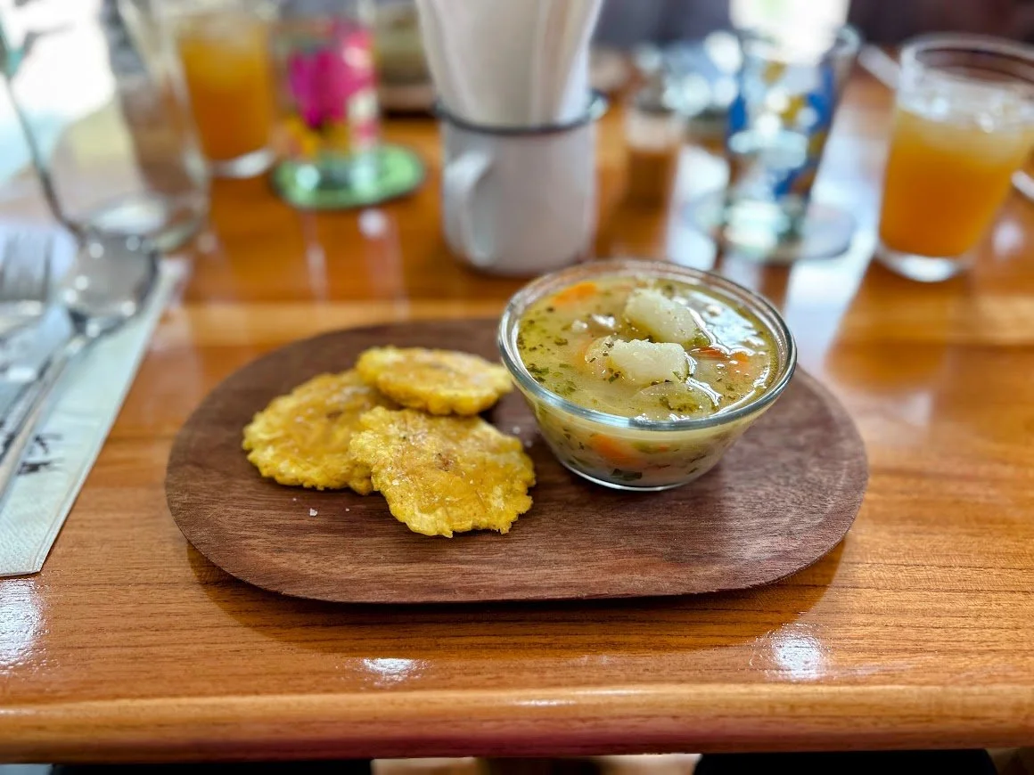 Fried plantains served with a bowl of vegetable soup on a wooden tray, set on a table with drinks and utensils in the background.
