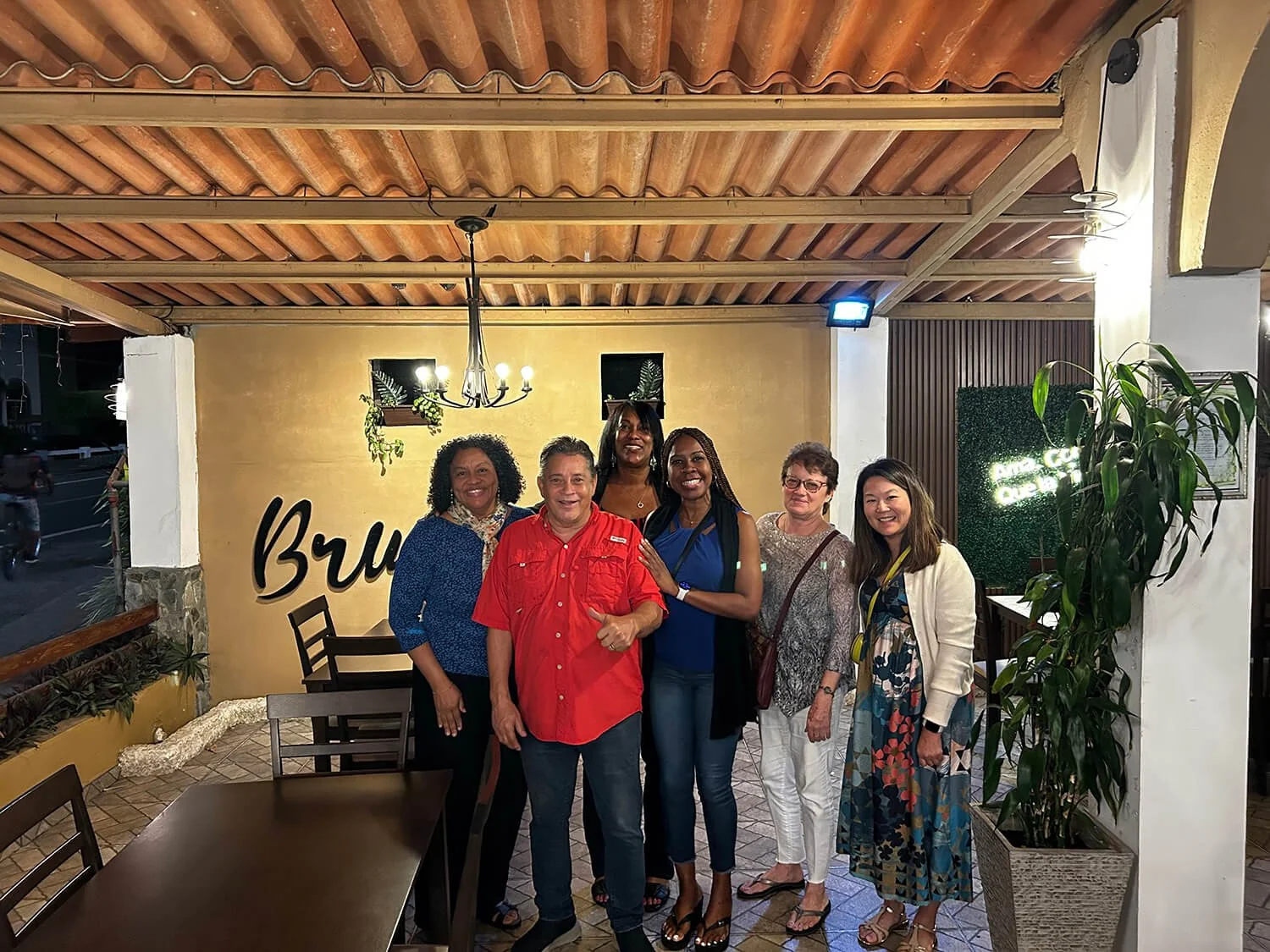 Group of six diverse people smiling at a restaurant or cafe, standing close together with some placing hands on each other's shoulders. The background features a yellow wall, a chandelier hanging from the wooden ceiling, potted plants, and a glowing 