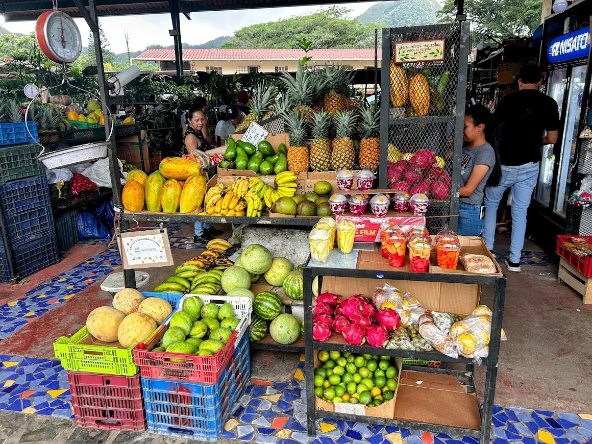 A fruit stand at an outdoor market with a variety of tropical fruits including pineapples, bananas, watermelons, papayas, and dragon fruits. There are some people shopping and a water dispenser on the right side of the image.