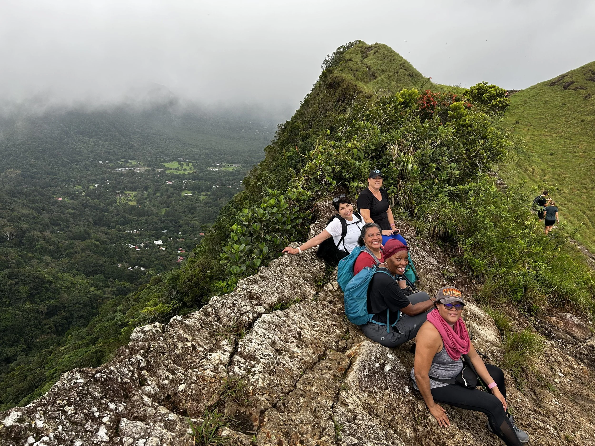 Group of hikers sitting on a rocky mountain trail with lush green scenery and a foggy mountain in the background