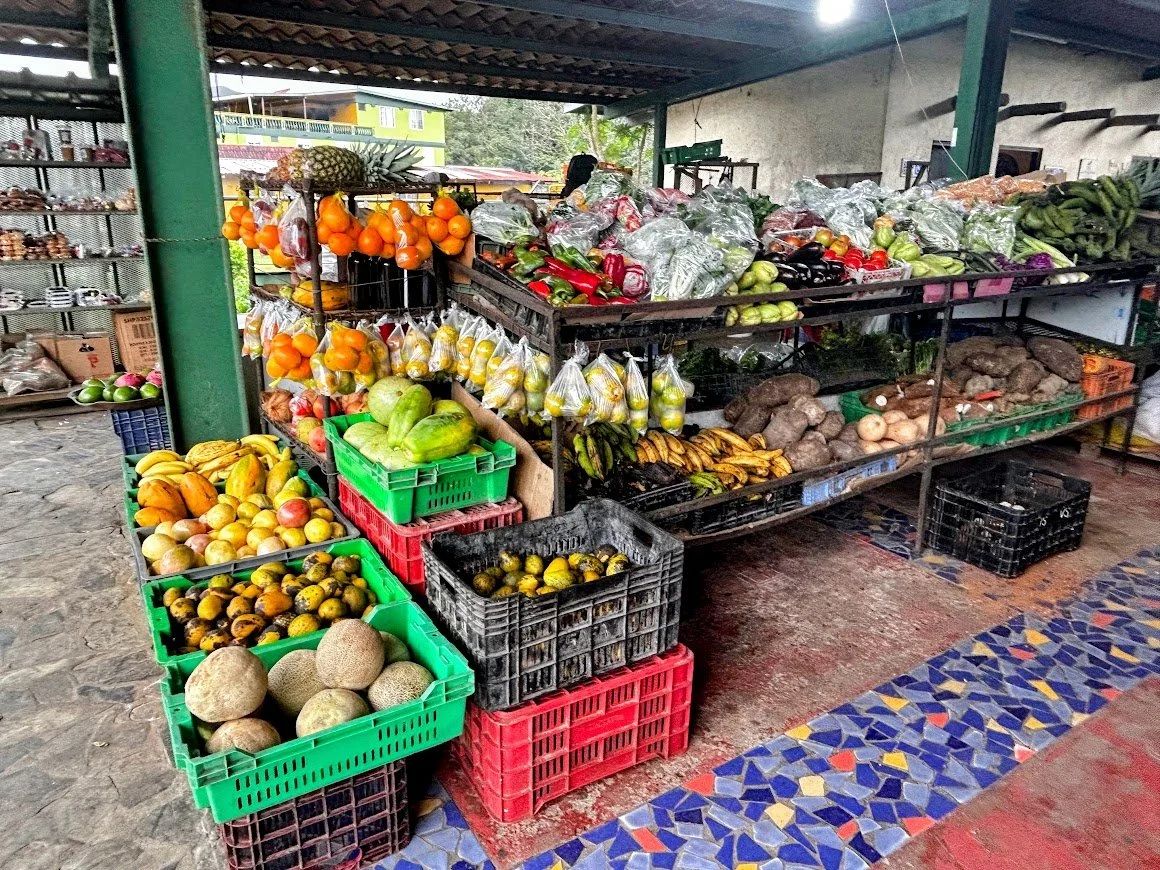 Fruits and vegetables on display at a market stand under a roof, including oranges, bananas, melons, pineapples, and various greens.