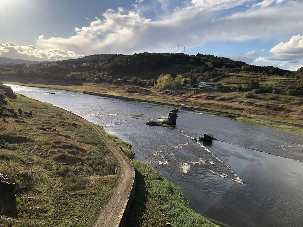 A wide river flowing through a hilly landscape with greenery, houses, and a partly cloudy sky overhead.