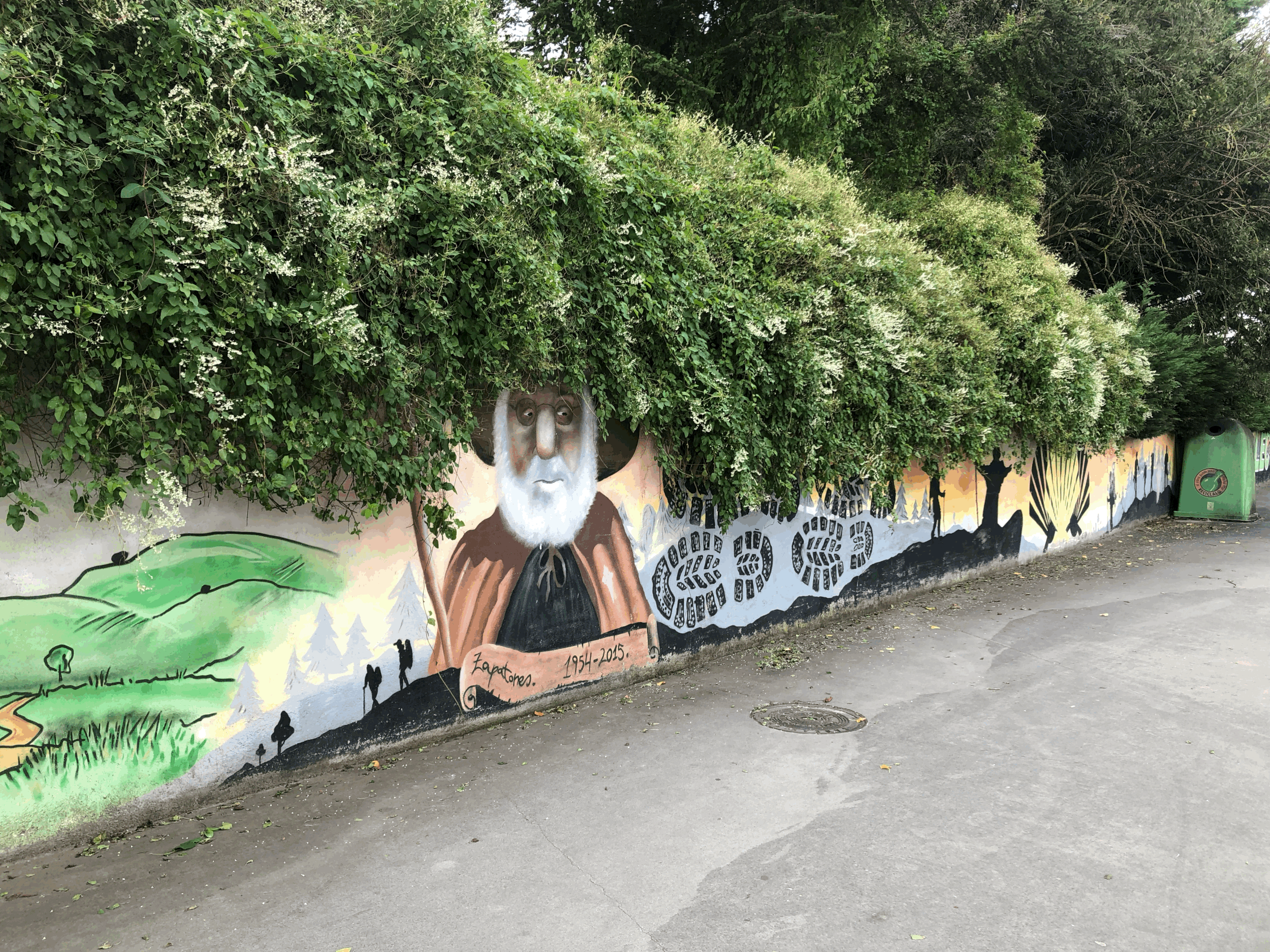 A mural painted on a wall depicting a person with a large white beard, wearing a hat, with a background of trees and nature. The mural also features black shoeprints, trees, and text reading 'Zapatones 1954-2015.'
