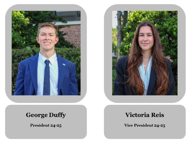 Two students, a young man and woman, standing outdoors in front of trees and a building. The young man is smiling and wearing a blue suit with a light blue shirt and blue tie, with the name tag 'George Duffy, President 24-25.' The young woman has long, wavy brown hair and is dressed in a dark blazer and light blue blouse, with the name tag 'Victoria Reis, Vice President 24-25.'