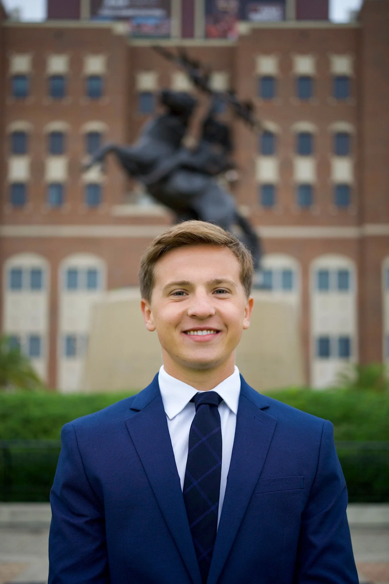 Young man in a blue suit and tie smiling in front of a statue of a horse with a rider on a building background.