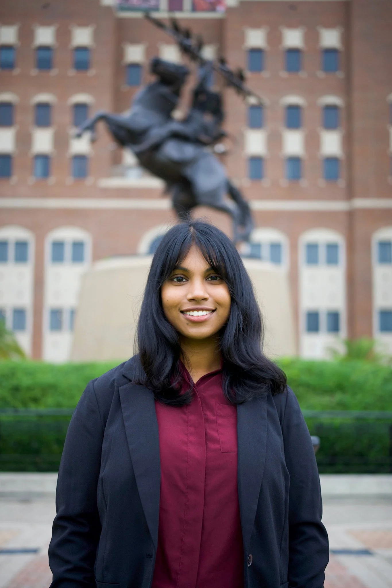 A young woman with dark hair smiling in front of a statue of a person on horseback, with a brick building in the background.