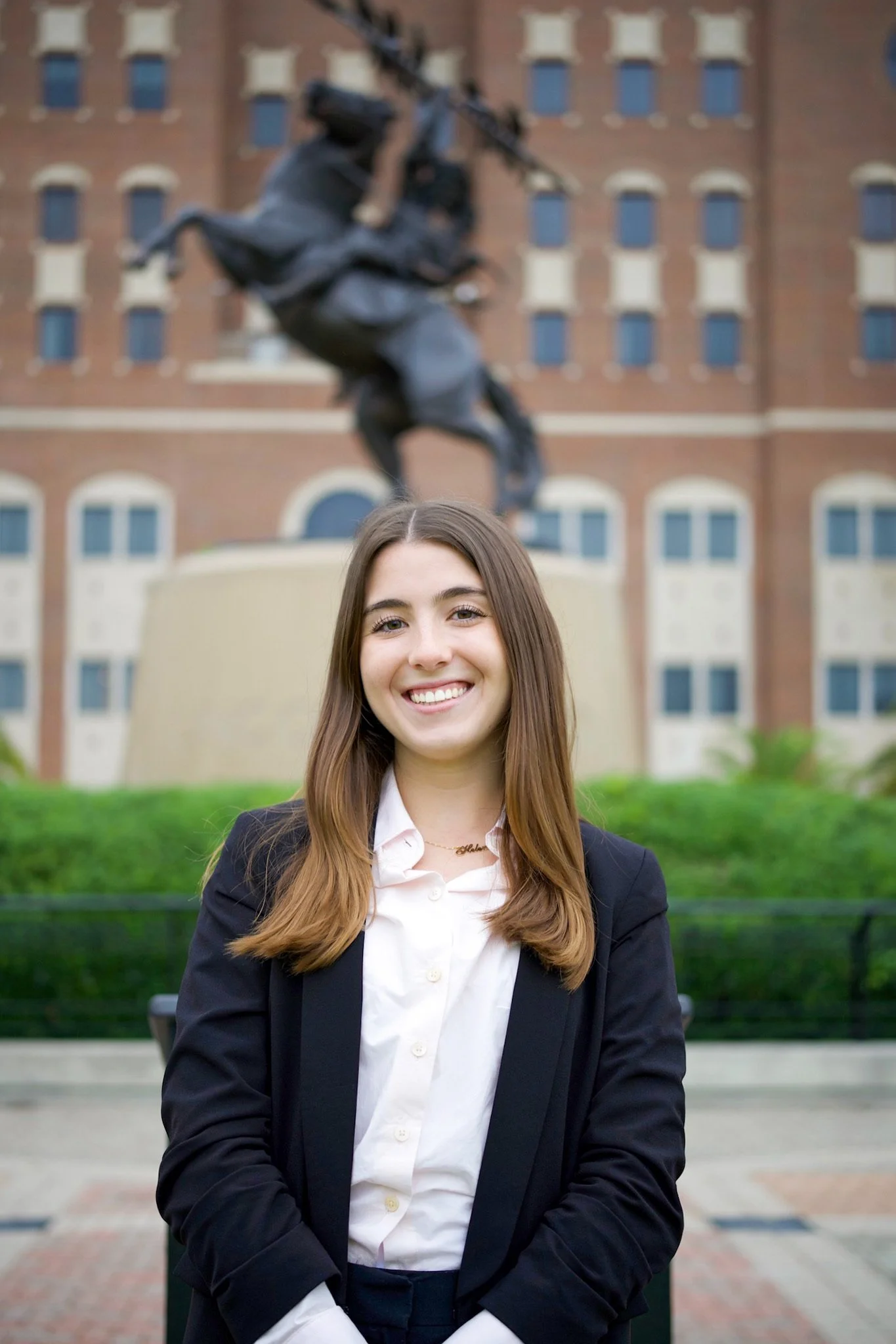 Young woman with long brown hair smiling in front of a statue of a horse and rider, on a university campus.
