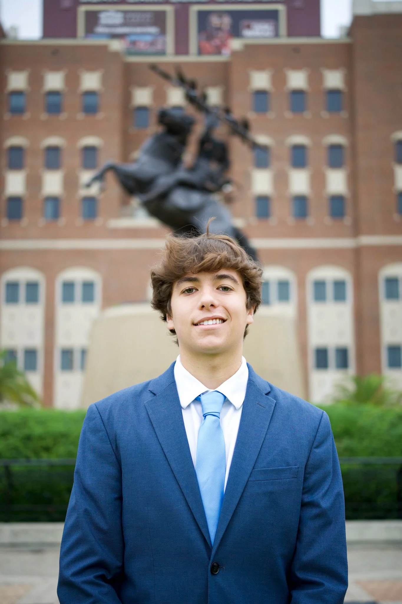 Young man in a blue suit and tie standing in front of a historic building with a statue of a soldier on horseback in the background.