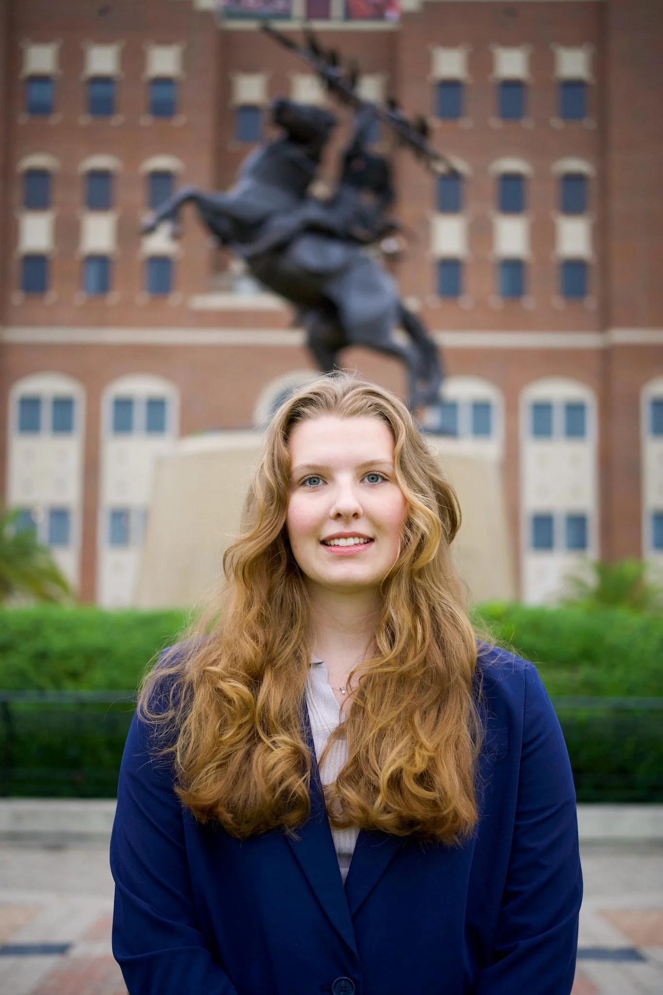 A woman with long, curly red hair, wearing a dark blue blazer, standing outdoors in front of a large building and a statue of a figure on a horse.