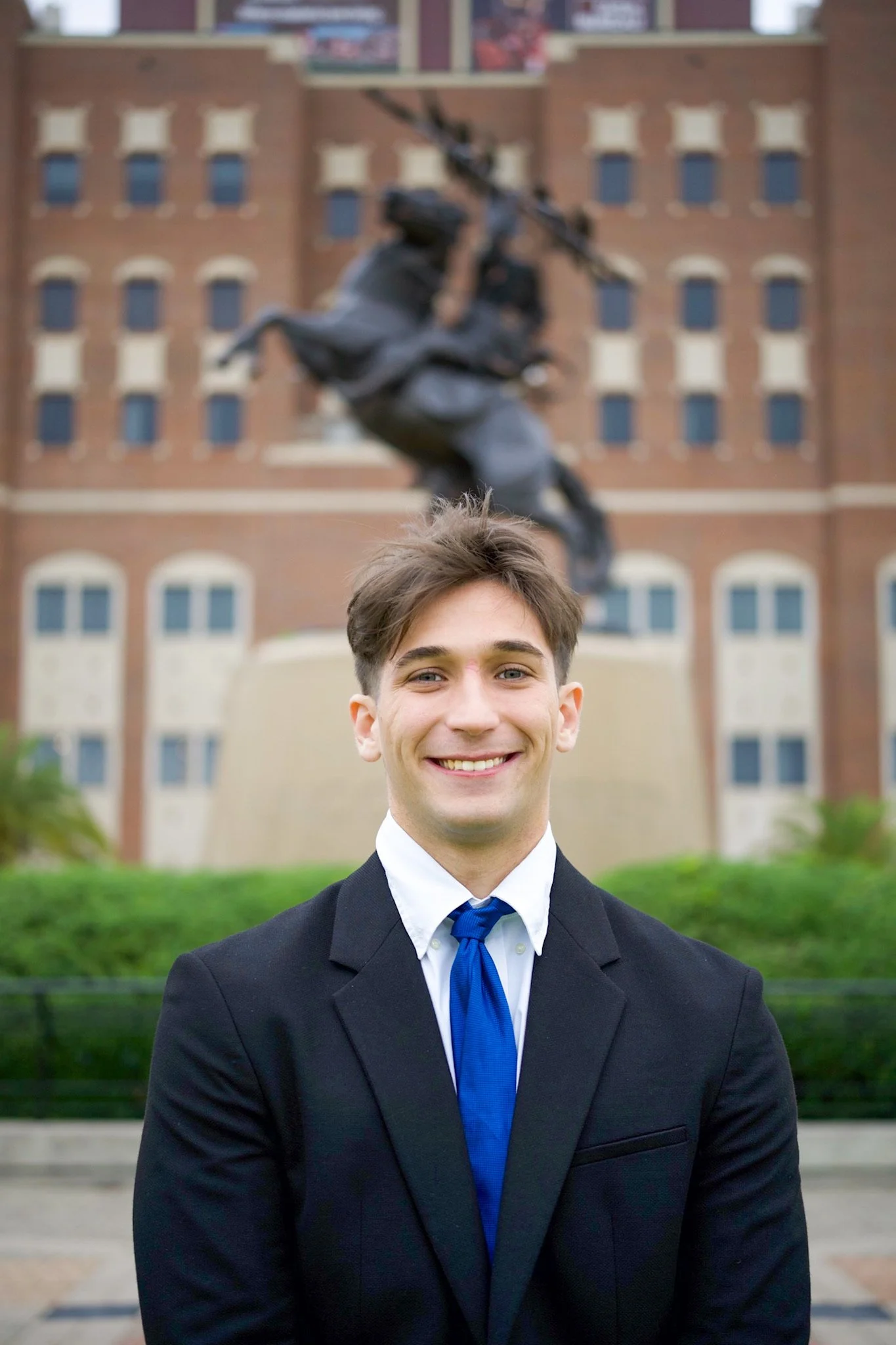 Young man in a suit smiling outdoors in front of a statue and a brick building.