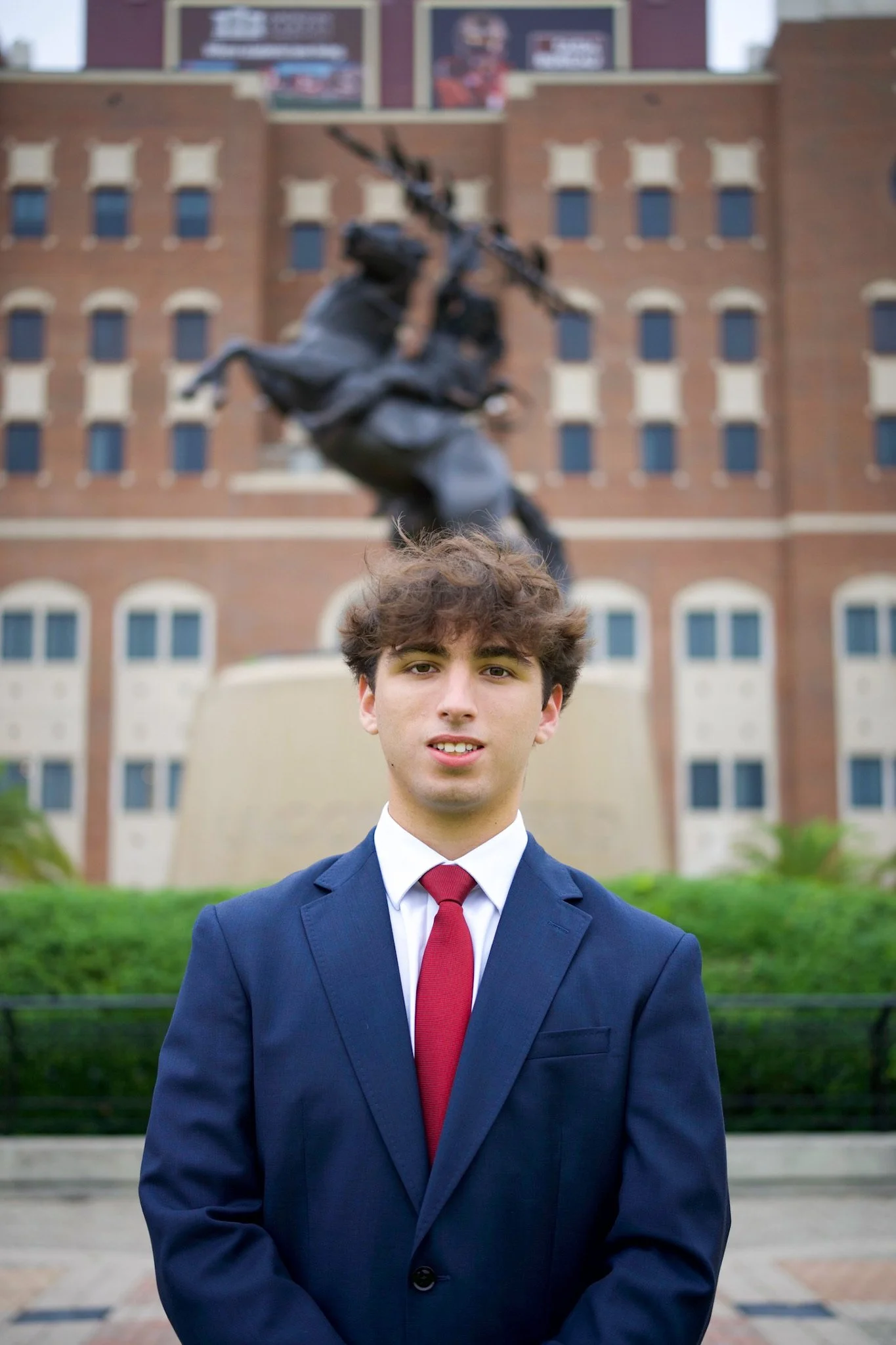 Young man in suit with red tie standing outdoors in front of a brick building and a statue.