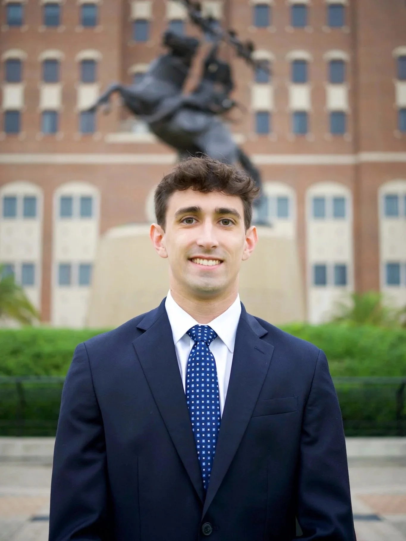 Young man in a navy suit and polka dot tie standing outdoors in front of a historic building and a statue.