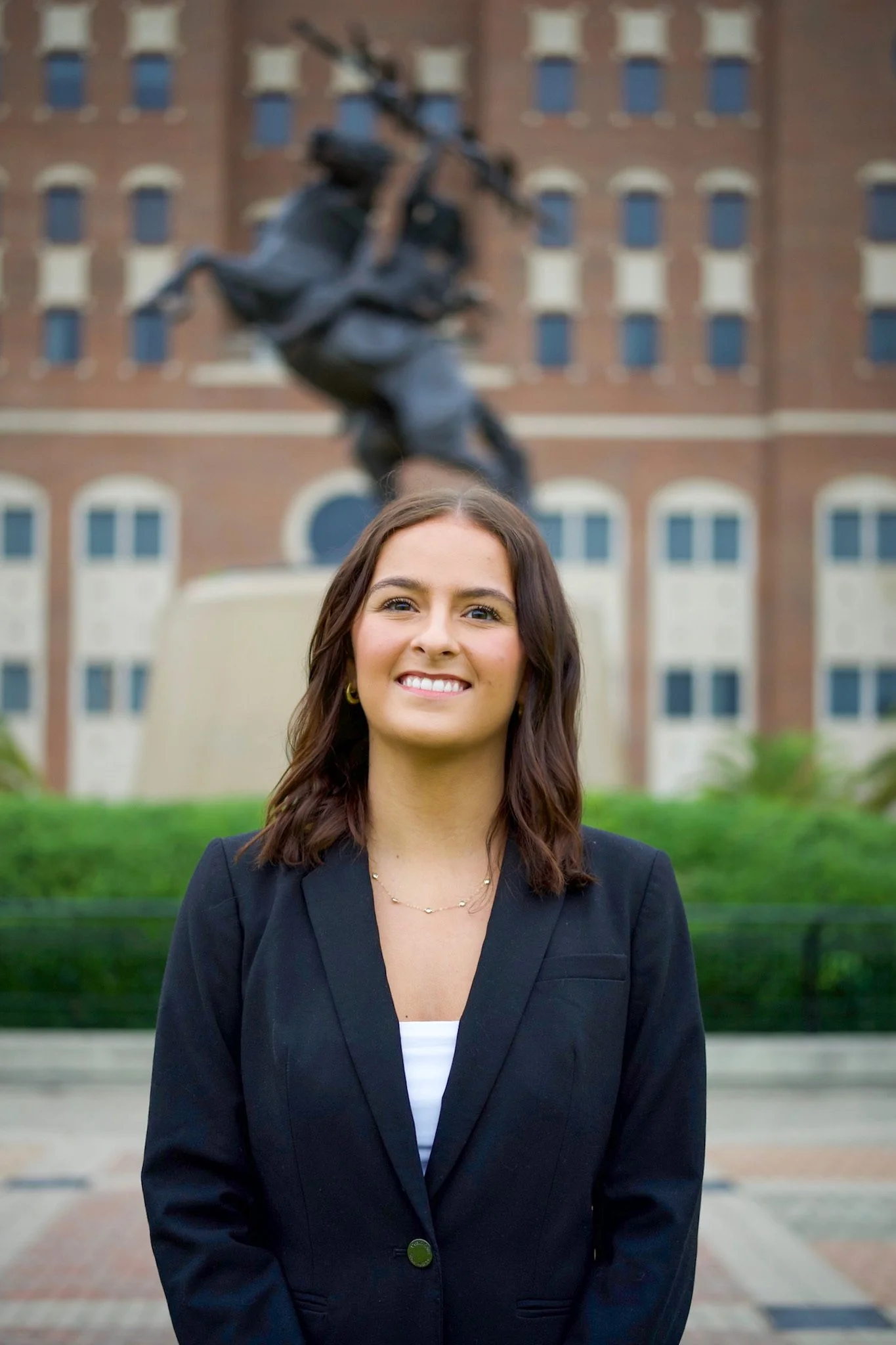 A young woman with shoulder-length brown hair, wearing a black blazer and a white shirt, smiling outdoors in front of a building with a statue of a rearing horse and rider in the background.