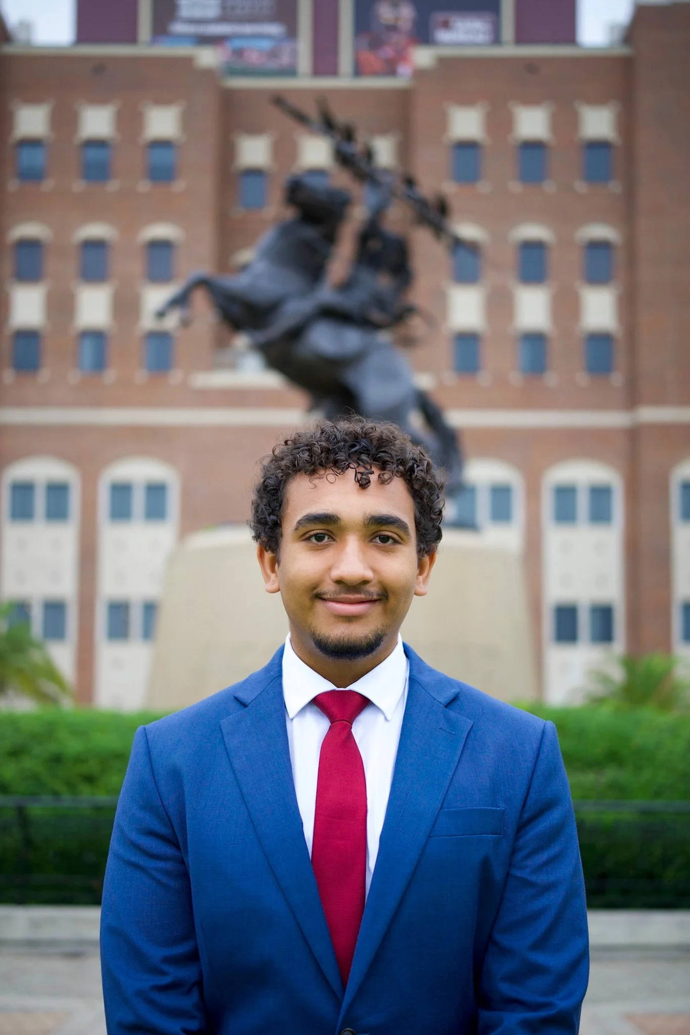 A young man in a blue suit with a white shirt and red tie standing in front of a bronze statue of a soldier with a rifle, and a brick building in the background.