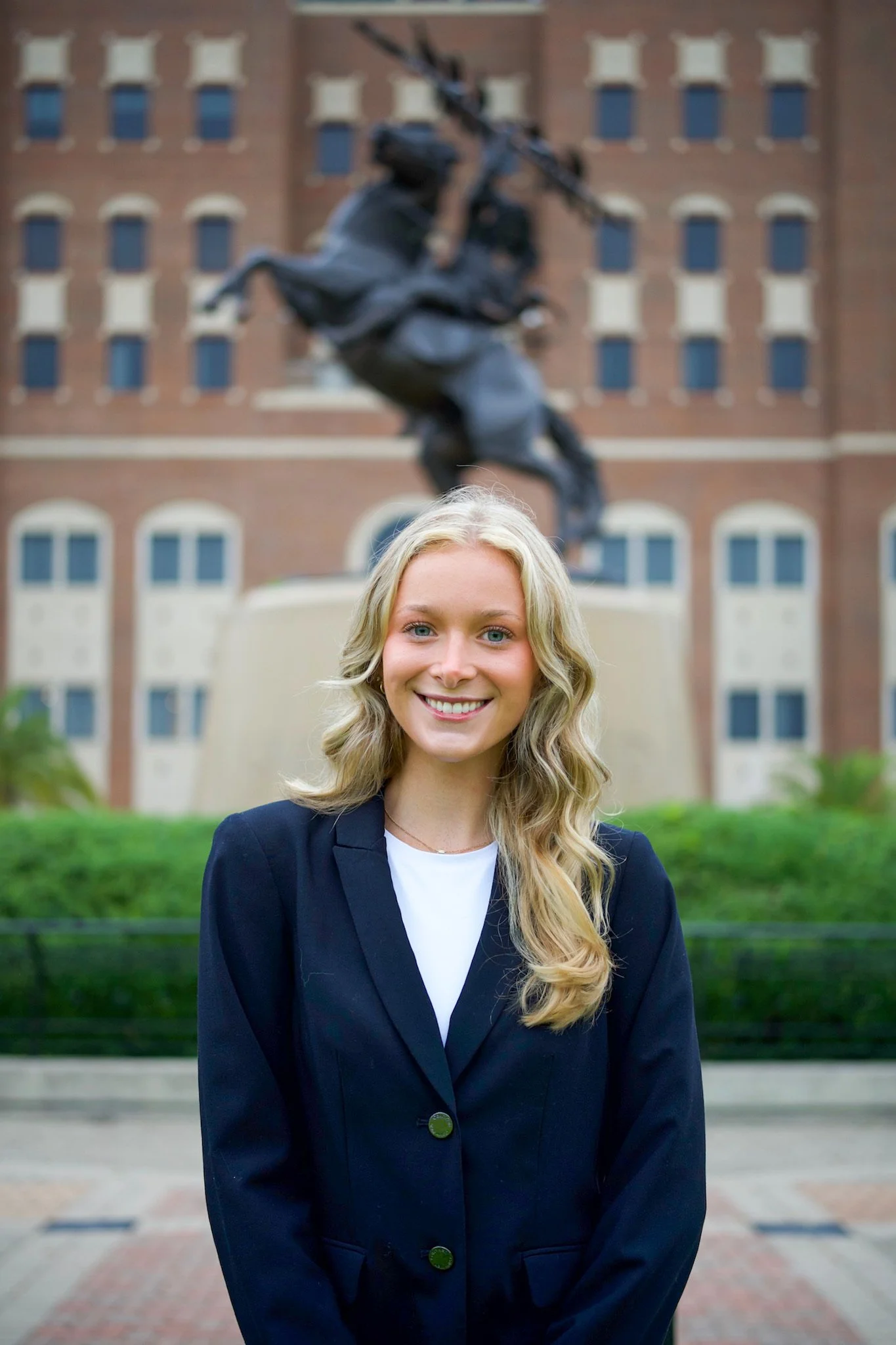 A woman in a navy blazer and white top smiling in front of a statue and historic brick building.