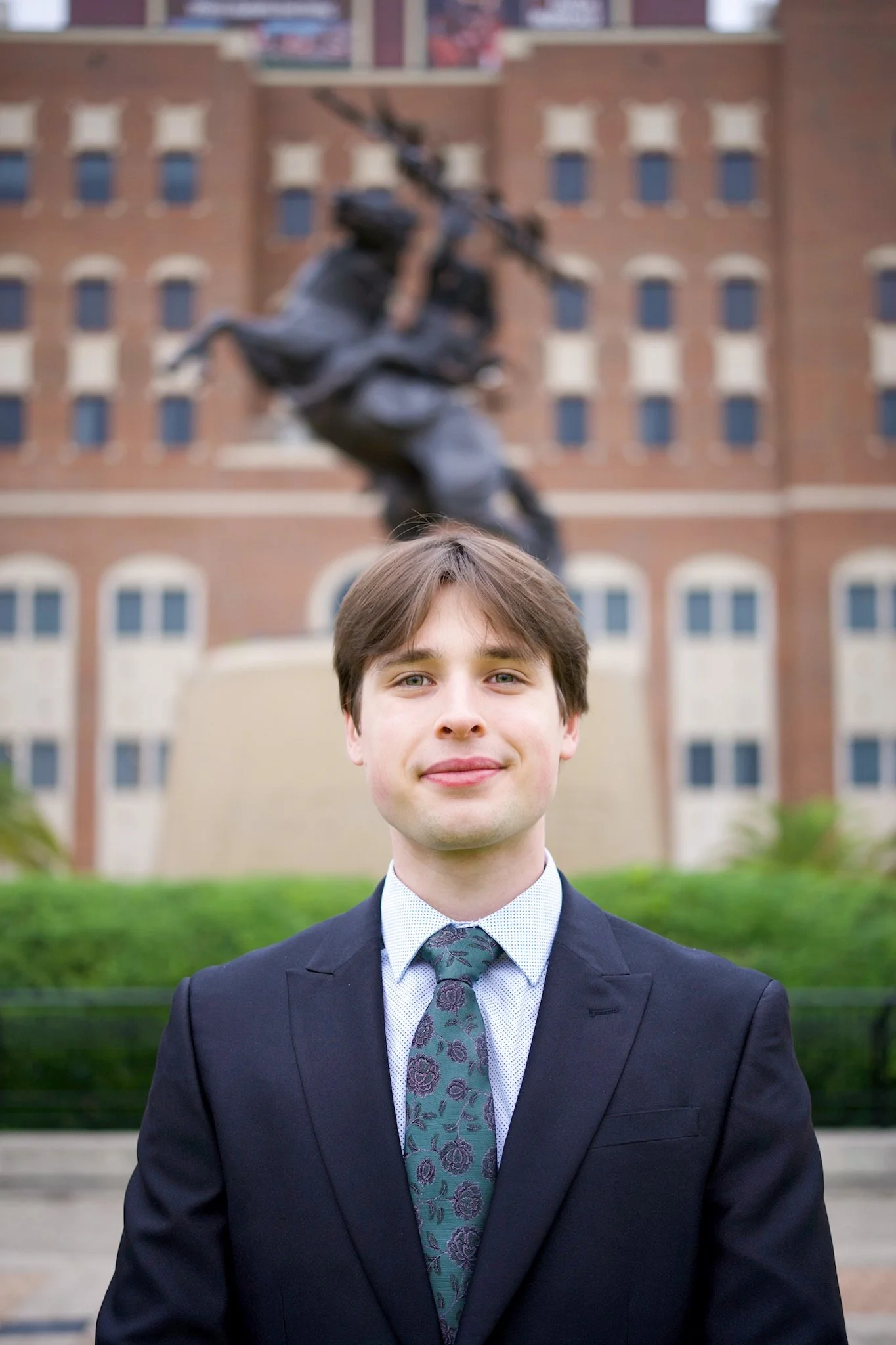 A young man in a suit and tie standing outdoors in front of a building with a statue of a soldier in the background.