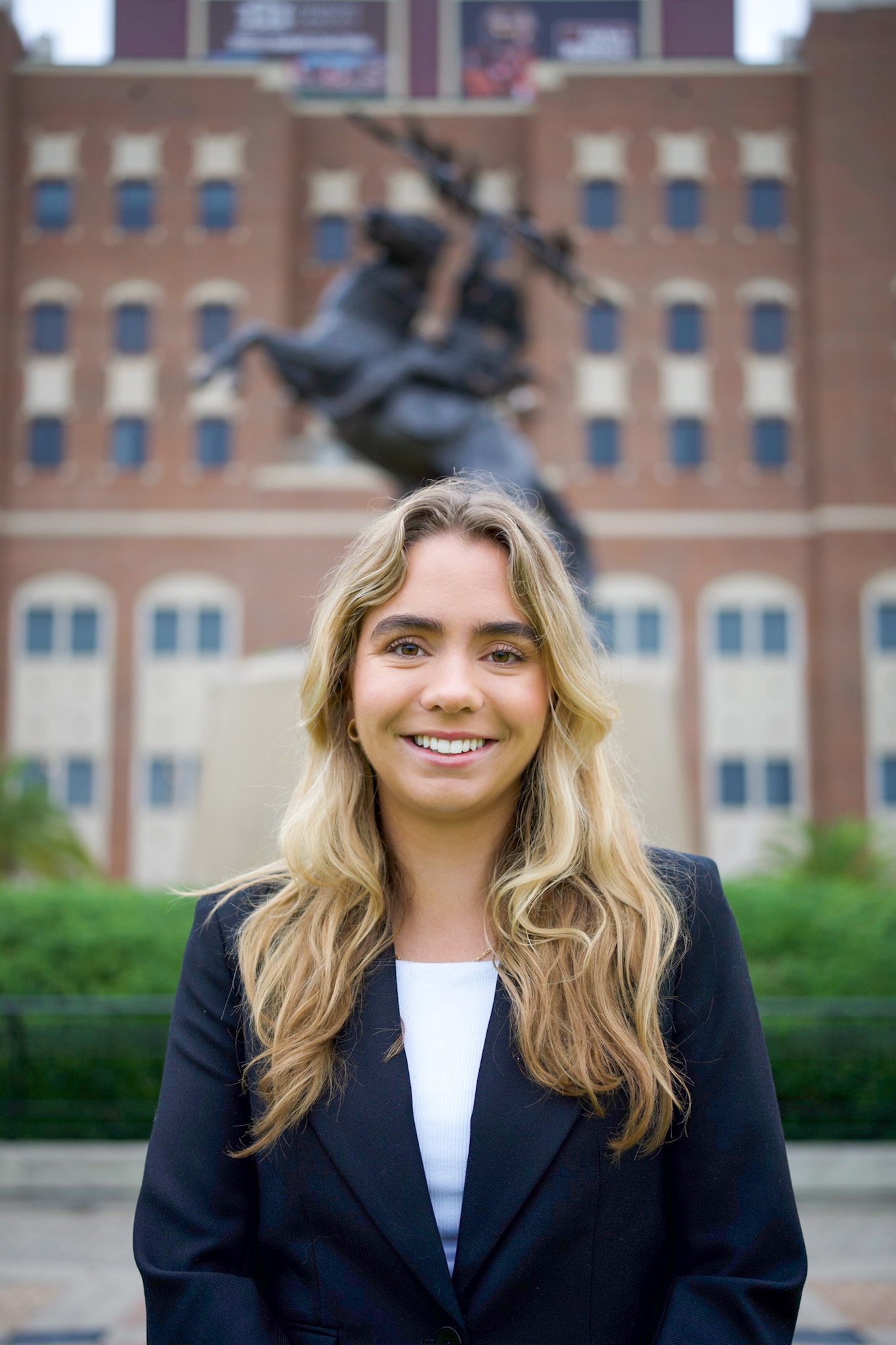 A smiling woman in professional attire standing outdoors in front of a monument with a statue of a warrior on horseback in the background, in front of a brick building.