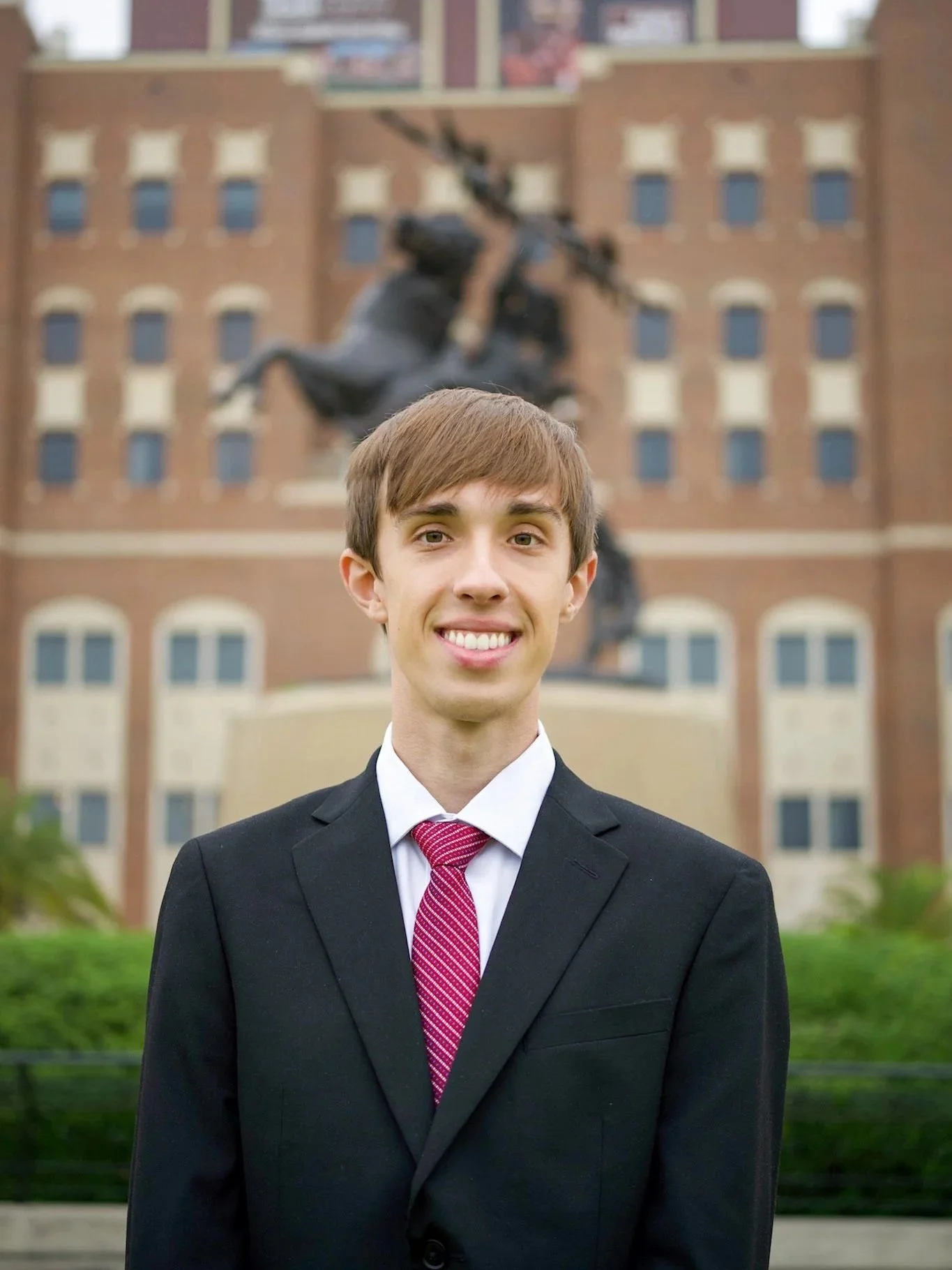 A young man in a black suit, white shirt, and red tie standing outdoors in front of a large brick building with a statue of an angel playing a harp in the background.
