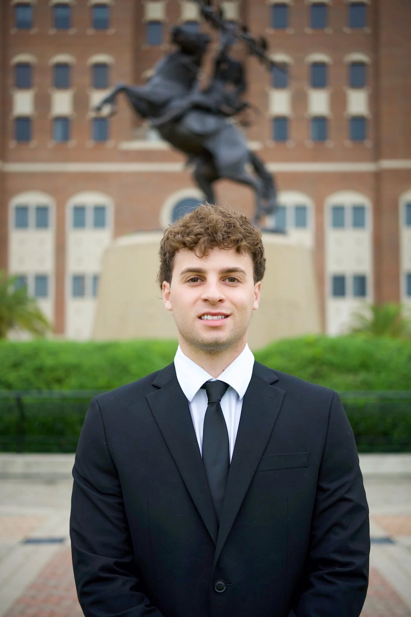 Young man in a black suit and tie standing outdoors, with a statue of a horse and rider in the background, in front of a brick building.
