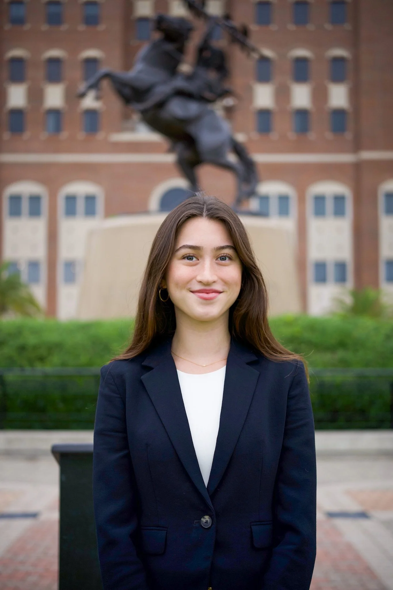 A woman smiling in front of a university building with a statue of a knight on horseback in the background.