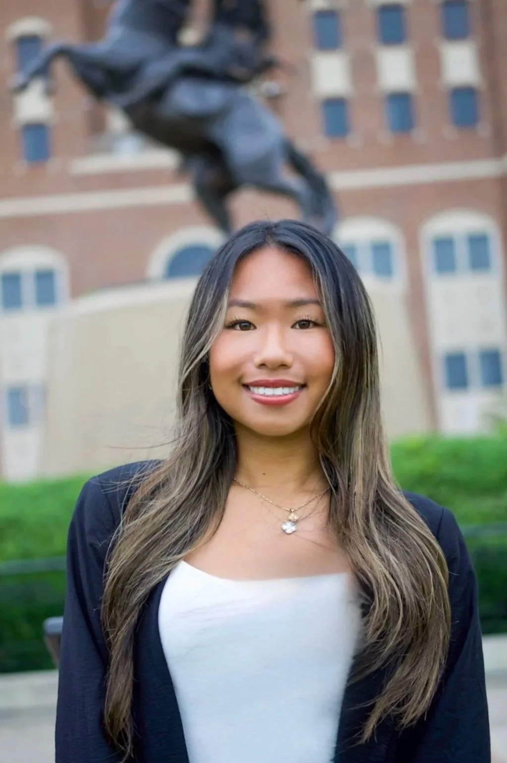A young woman smiling outdoors in front of a large building and a statue of a horse and rider.