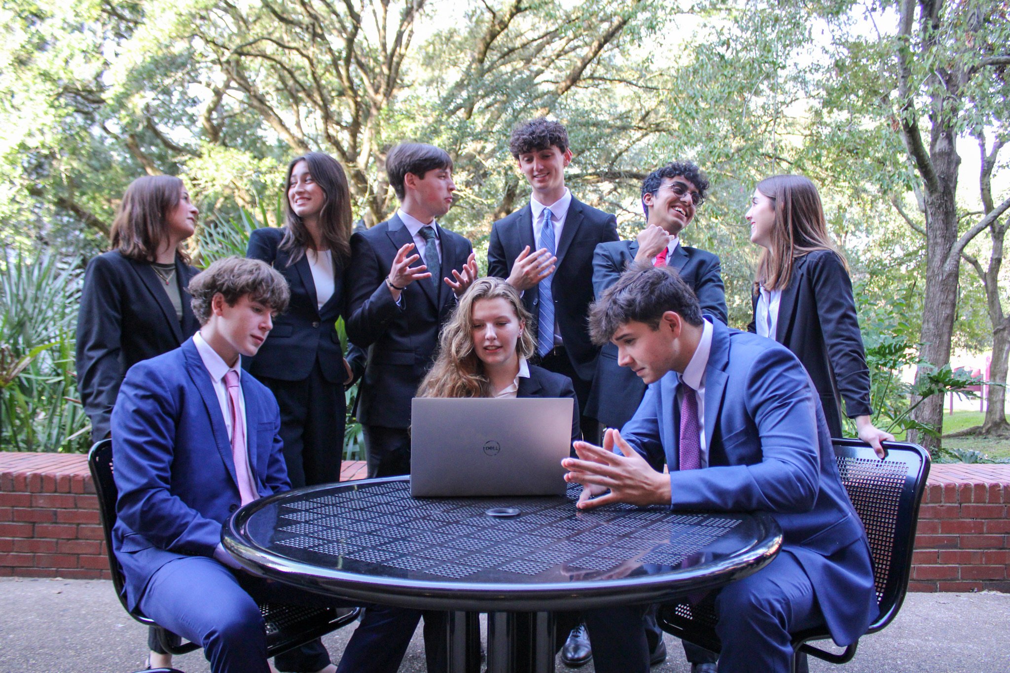 Group of young professionals in business attire gathered around a laptop outdoors, engaging in discussion and smiling.