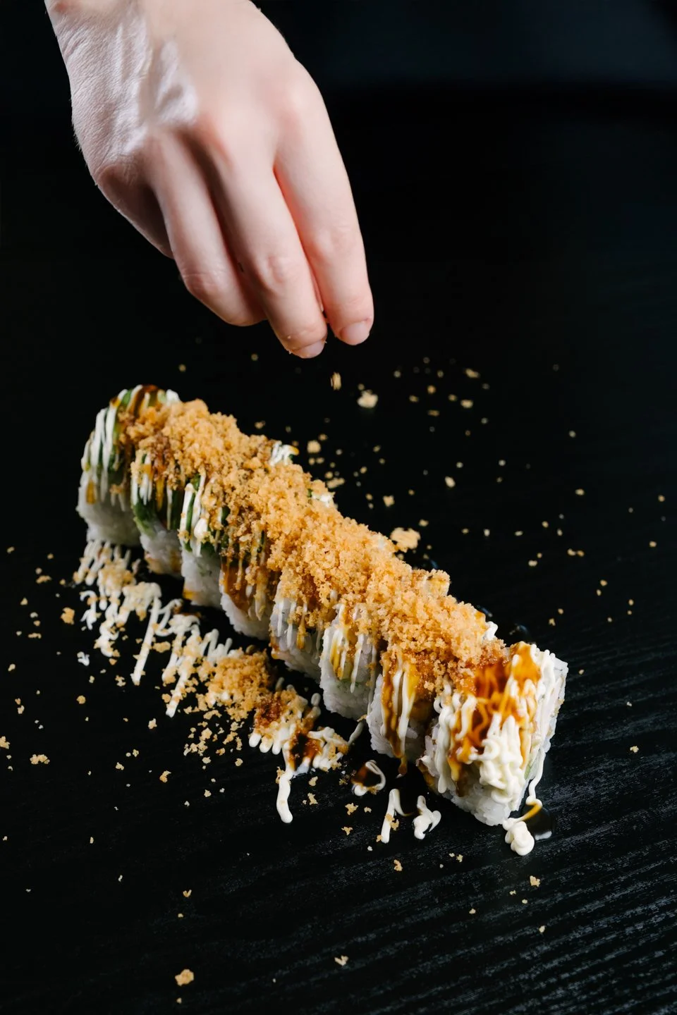 A hand sprinkling crushed topping on a sushi roll with cream cheese, drizzled with sauce and garnished with crumbs, on a black surface.