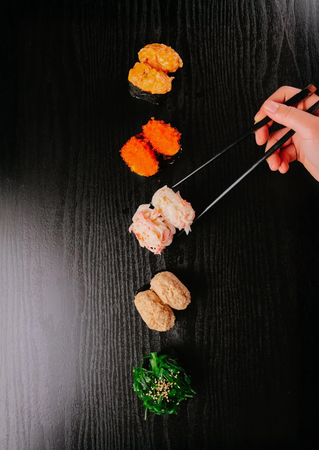 Assorted sushi pieces on a black surface, with a hand holding chopsticks picking up one piece of sushi topped with seafood.