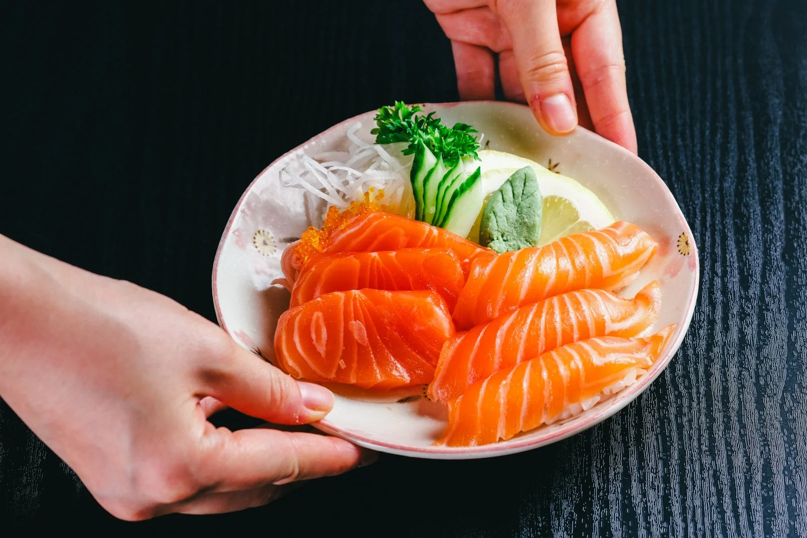 A person holding a bowl of assorted sashimi, including slices of salmon, garnished with wasabi, cucumber, lemon, and green parsley, on a dark wooden surface.