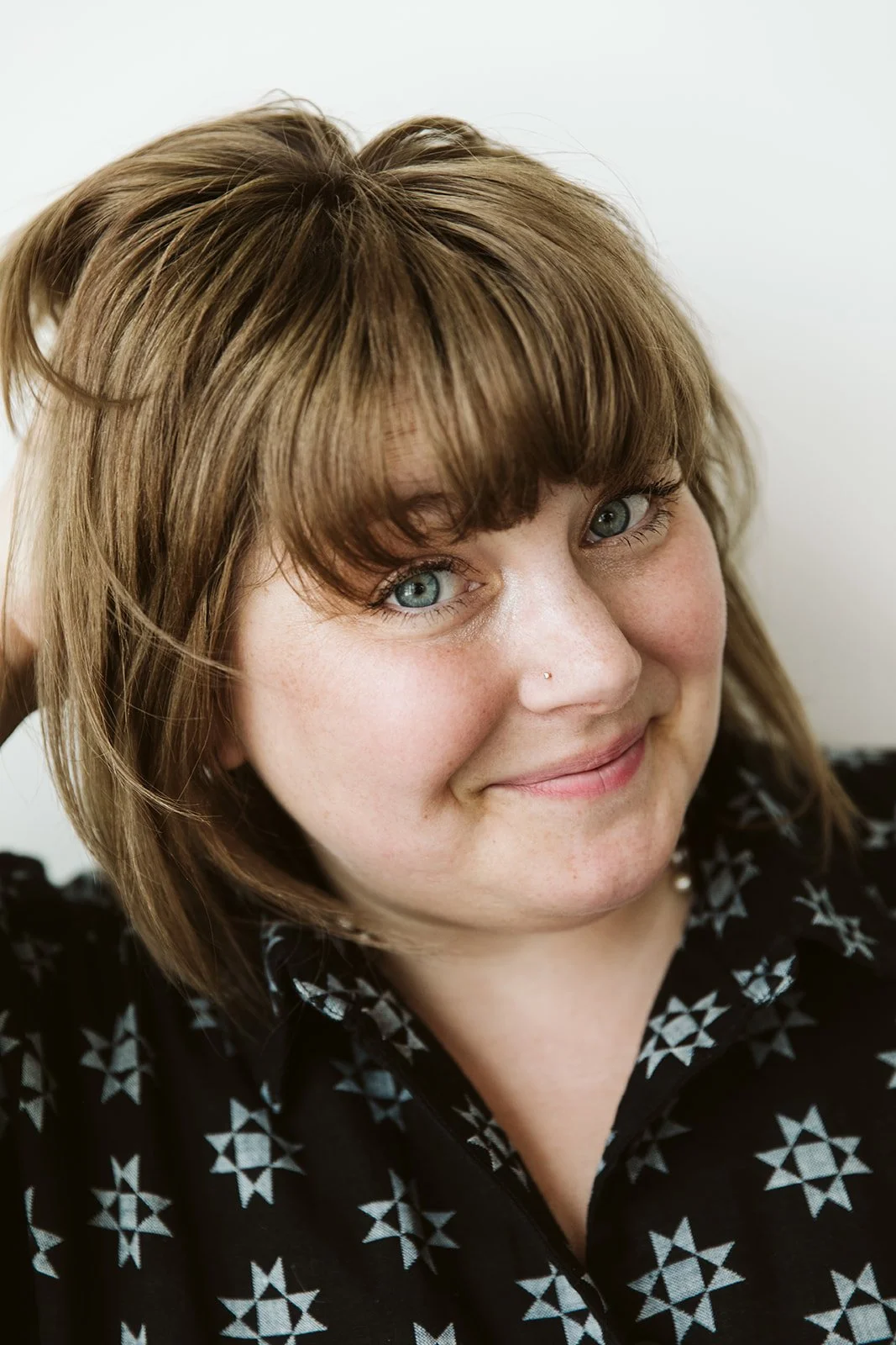 Close-up of a smiling woman with blue eyes, light brown hair with bangs, wearing a black top with white star patterns.